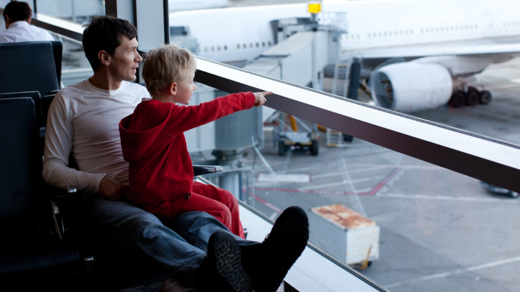 Son sits on dads lap while they look outside the window at airplanes on the tarmac